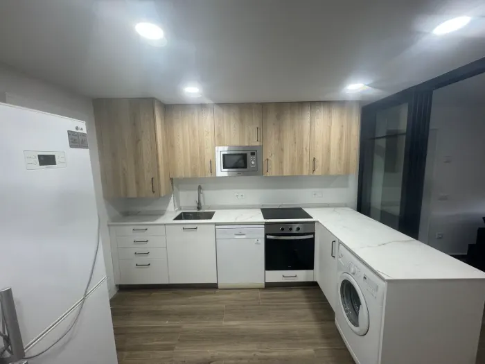 Cocina en L con muebles bajos blancos, altos en madera clara y encimera blanca